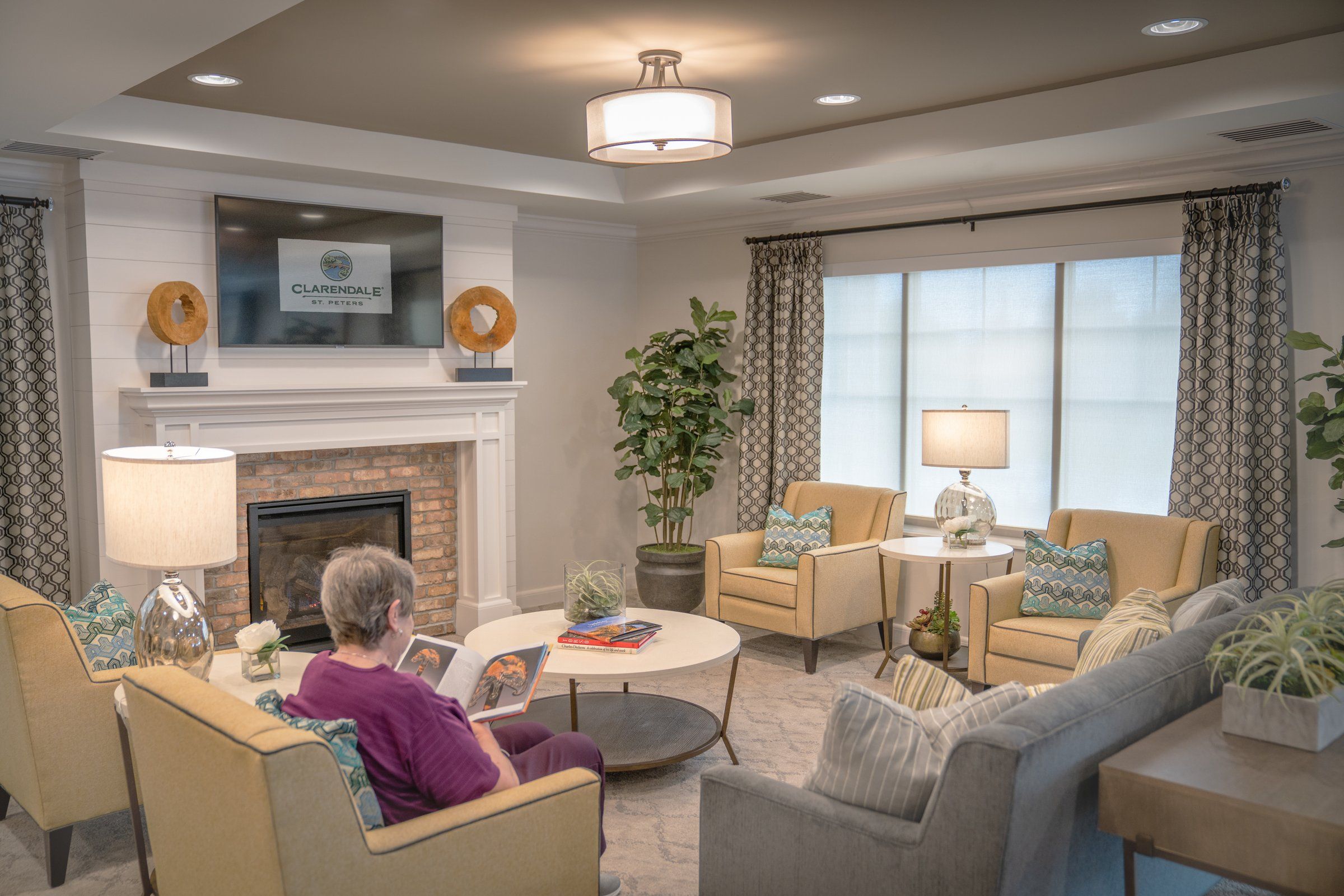 A resident reads in a lounge with a brick fireplace, plants and warm lamps