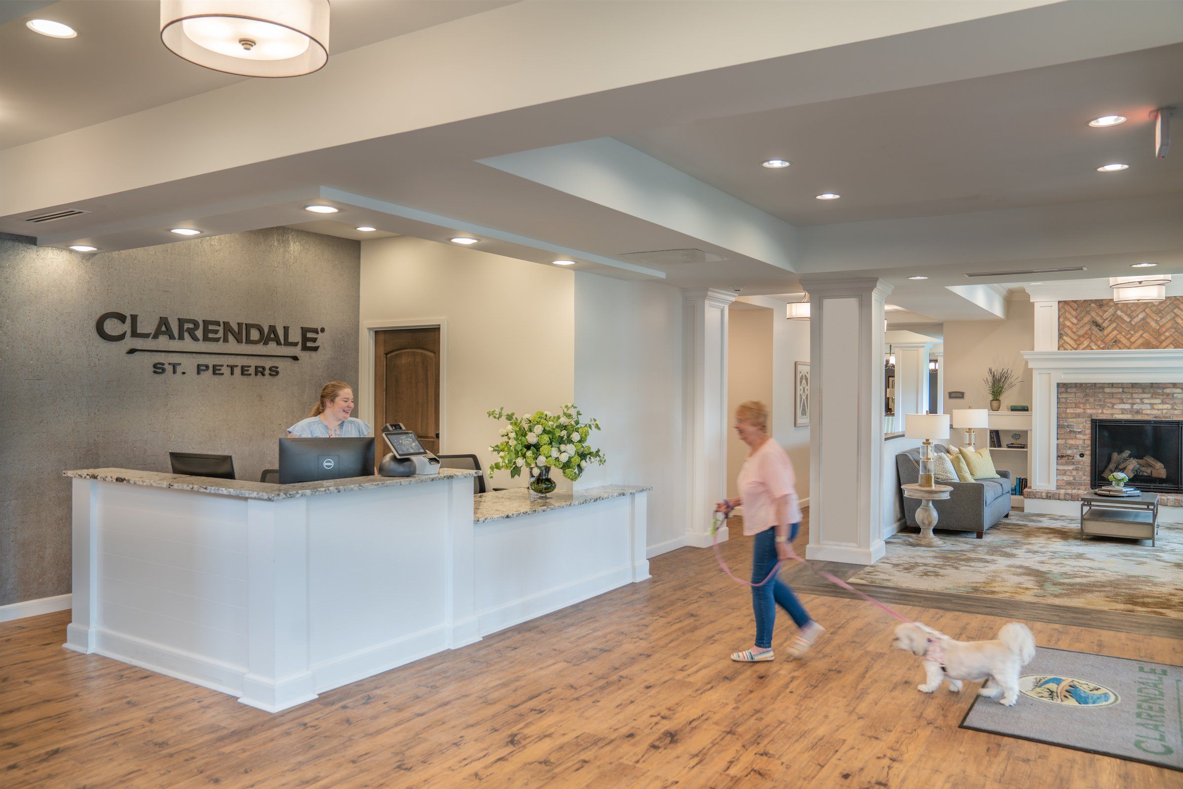 A staff member at the reception desk and a resident walking a small dog across a warm, wood-floored lobby