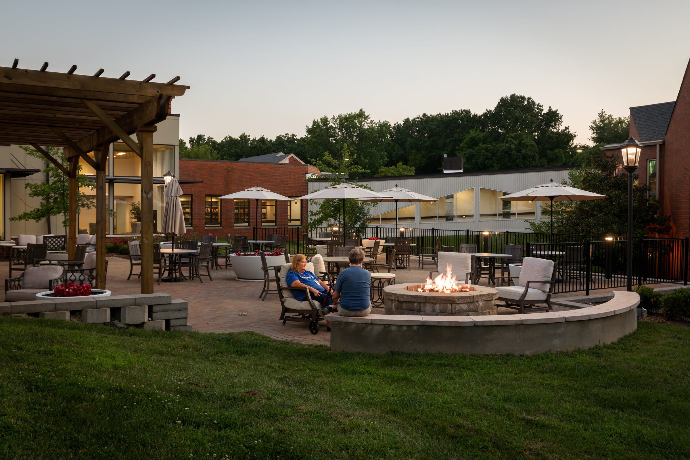 Two residents sit by a fire pit in a courtyard with a pergola and umbrellas at dusk