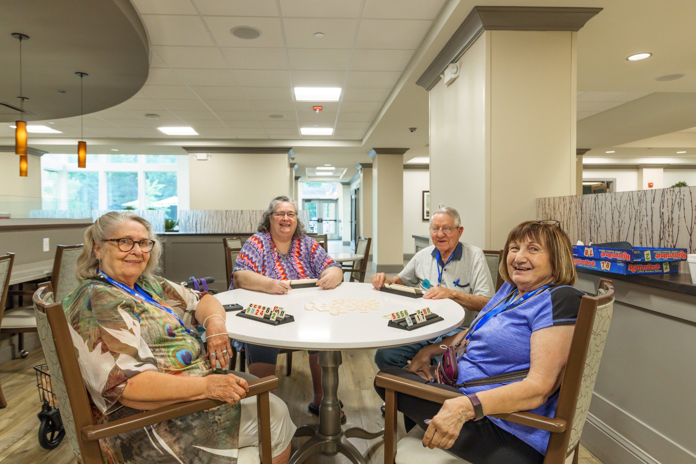 Four residents smile around a table while playing a tile game in a bright community room