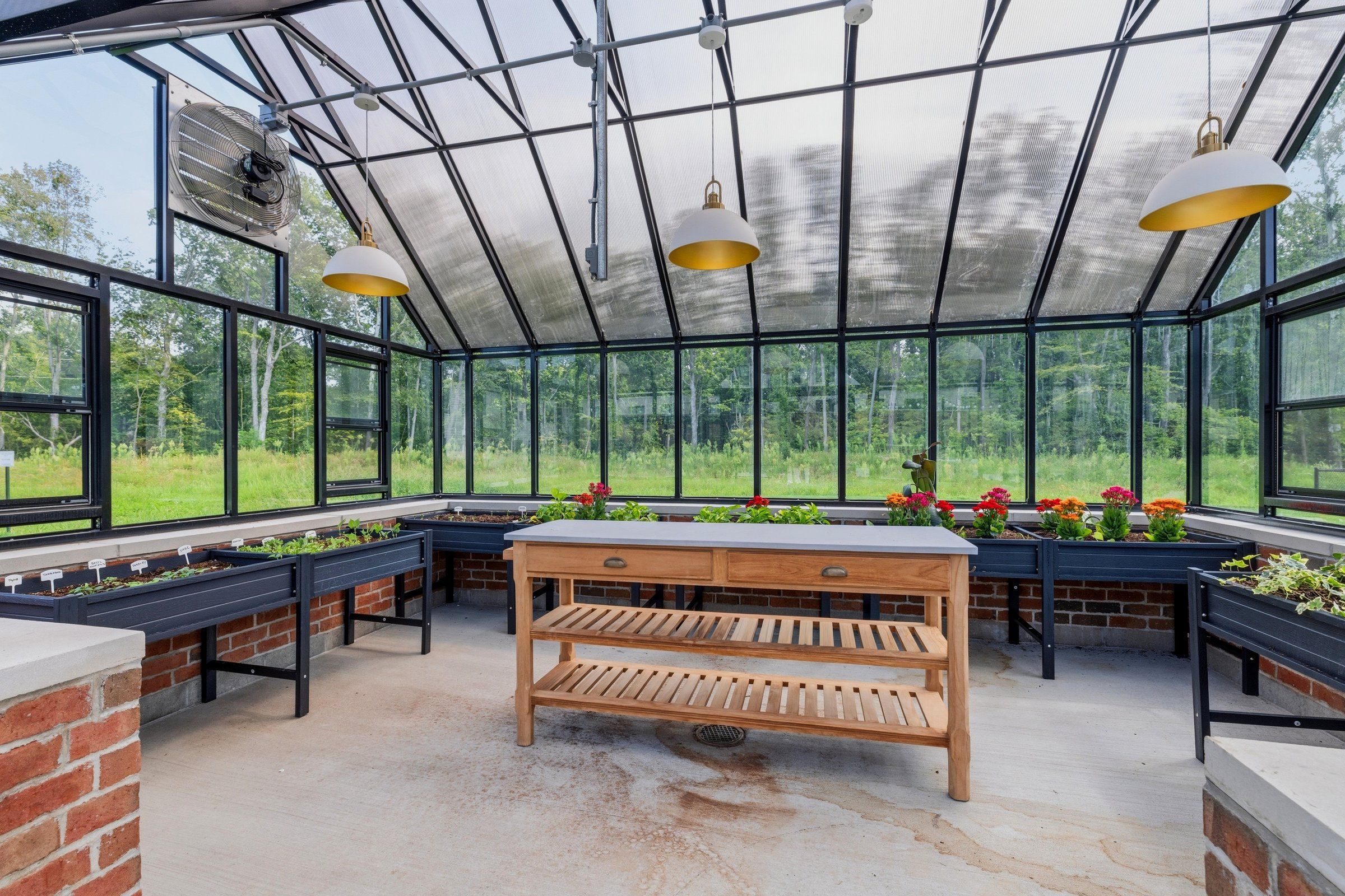 A glass greenhouse with raised planting beds, a wood work table and trees beyond
