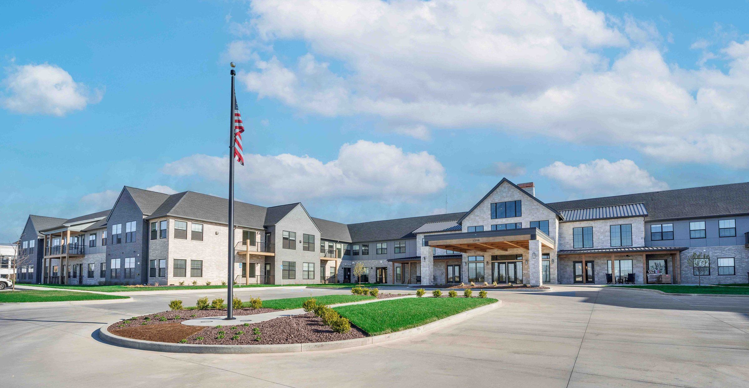 Cedarhurst of Wentzville, a building of gables, stone and siding with a covered entry and a flag, seen across the drive
